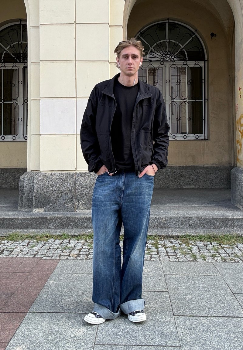 Giovane uomo con capelli biondi, giacca nera, jeans larghi blu e sneakers, in piedi con le mani nelle tasche davanti a un ingresso arcuato di un edificio.