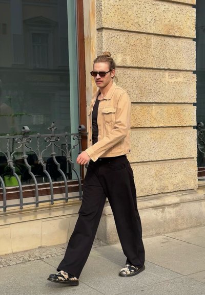 Chaqueta de mezclilla beige sobre una camisa negra, pantalones anchos negros y zapatos con estampado. Las gafas de sol completan el conjunto. Muro de piedra al fondo.