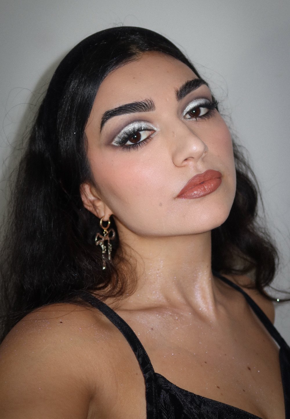 Nærbilde av en person med en svart topp med blomsterstruktur, sølvøyenskygge og gullkors øreringer, med glitter på huden.