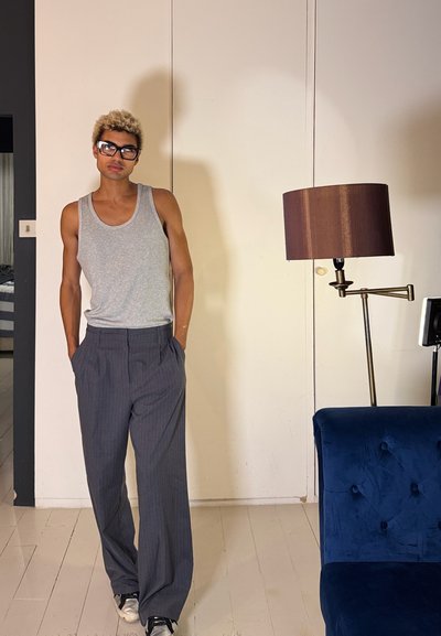 Persona con cabello corto, rubio y rizado, con gafas, vistiendo una camiseta sin mangas gris y pantalones de rayas, de pie en el interior cerca de una silla azul y una lámpara de pie.