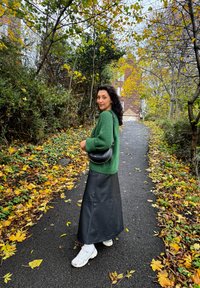 Green knitted sweater, black leather maxi skirt, white sneakers, and a black handbag with studs, on a path lined with autumn leaves.