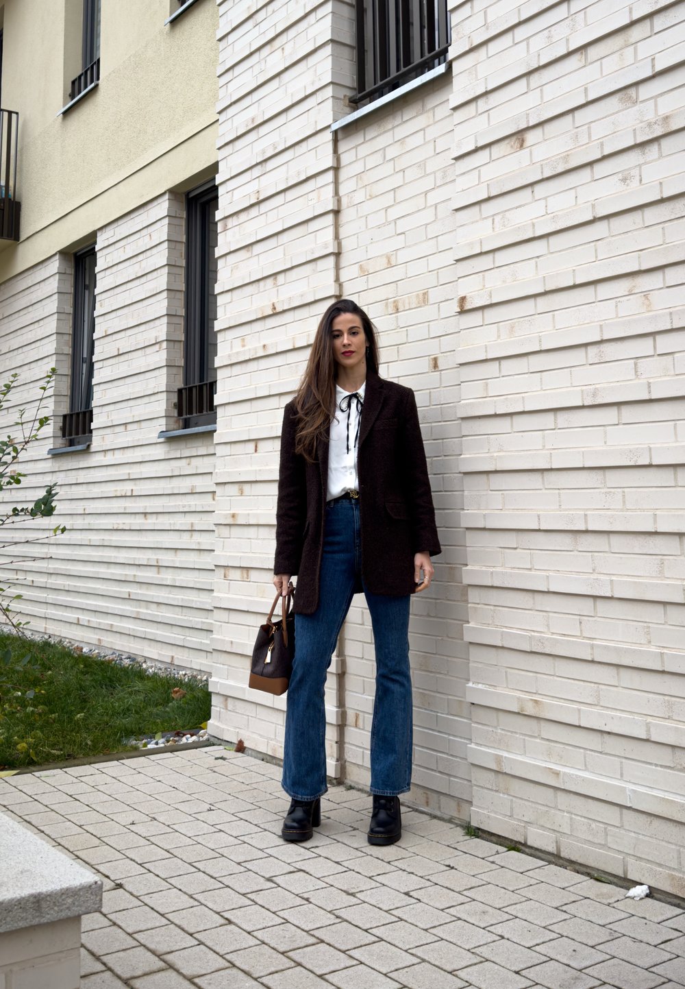 Femme debout près d'un mur de briques blanches, portant un manteau noir, un chemisier blanc, un jean bleu, des bottes noires, tenant un sac à main marron en extérieur.