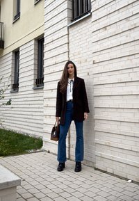 Femme debout près d'un mur de briques blanches, portant un manteau noir, un chemisier blanc, un jean bleu, des bottes noires, tenant un sac à main marron en extérieur.