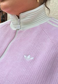 Gaiši rozā ribota Adidas džemperis ar baltu rāvējslēdzēja apkakli, uz krūtīm mazs balts izšūts logo un baltas svītras akcentiem.