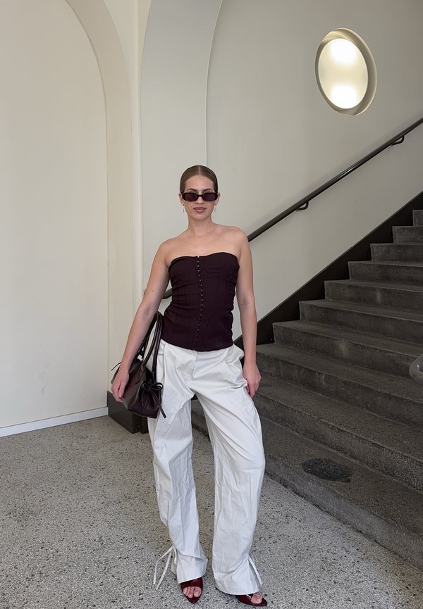 Femme portant un haut bustier foncé, un pantalon large blanc, des lunettes de soleil, tenant un sac à main foncé, debout près d'un escalier gris avec un mur courbé et une lumière ronde.