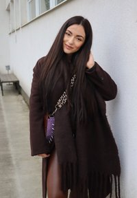 Rjavi volneni plašč z detajlom resic, kombiniran s vijoličasto tkano torbico z srebrnimi konicami. Ozadje je betonsko okolje.