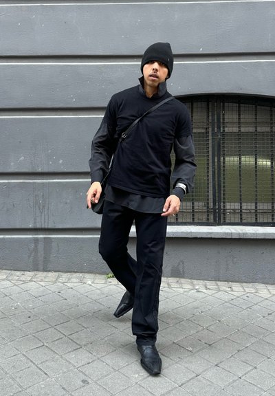 Hombre con gorro negro, camisa de manga larga en capas, pantalones y zapatos puntiagudos caminando por la acera de una ciudad junto a una pared gris con ventana con rejas.