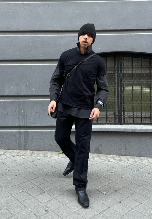 Homme portant un bonnet noir, une chemise à manches longues superposées, un pantalon et des chaussures pointues, marchant sur un trottoir en ville devant un mur gris avec une fenêtre grillagée.