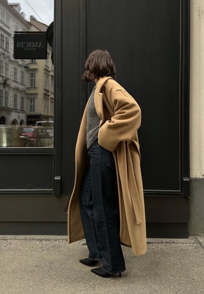 Abrigo largo de camel, suéter gris, jeans oscuros y zapatos negros. El abrigo presenta una cintura ceñida con cinturón y mangas amplias. Diseño simple y moderno.