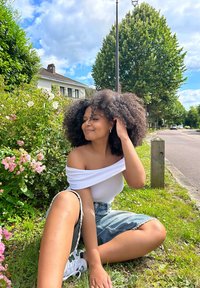 Persona seduta sull'erba, che indossa una maglietta bianca senza spalle e pantaloni shorts di jeans strappati. Capelli ricci, scarpe da ginnastica bianche, circondata da fiori rosa e vegetazione.