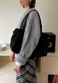 Bolso tote de ante negro con textura suave, con detalle de hebilla y asas para llevar. Combinado con un suéter gris de punto y una falda de cuadros.