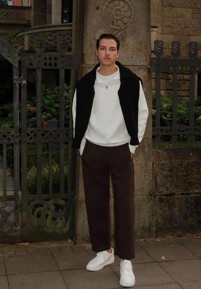 Sudadera blanca con cremallera sobre un chaleco sin mangas negro, pantalones marrones y zapatillas blancas. De pie contra una pared de piedra y una puerta.