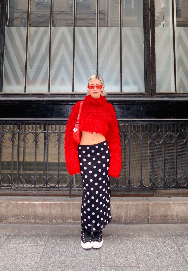 Osoba stojící venku, oblečená v červeném chlupatém krátkém svetru, červených slunečních brýlích, černobílé suknu s puntíky, černých teniskách a nesoucí červenou tašku.