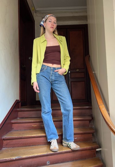 Chaqueta verde lima, top marrón corto, jeans rectos azules y zapatillas beige con acentos verdes, de pie en escaleras de madera.