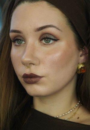 Make-up-Look mit glanzvollen braunen Lippen, definiertem Eyeliner und schimmerndem Lidschatten. Ein goldener Septumring und Ohrringe setzen Akzente. Braunes Kopfband.