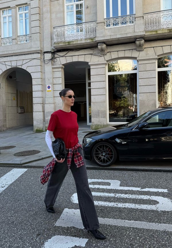 Mujer con camisa roja y gafas de sol cruza una calle frente a un coche negro y un edificio de piedra con grandes ventanas.