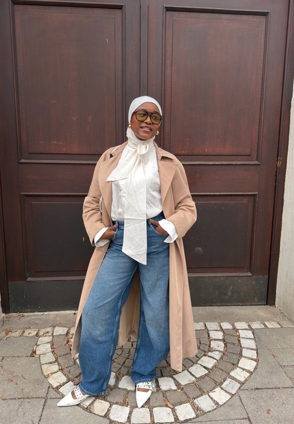 Kvinde iført beige trenchcoat, hvid bluse med sløjfe, blå viddebenede jeans, hvide spidse sko og tørklæde står foran store trædøre.