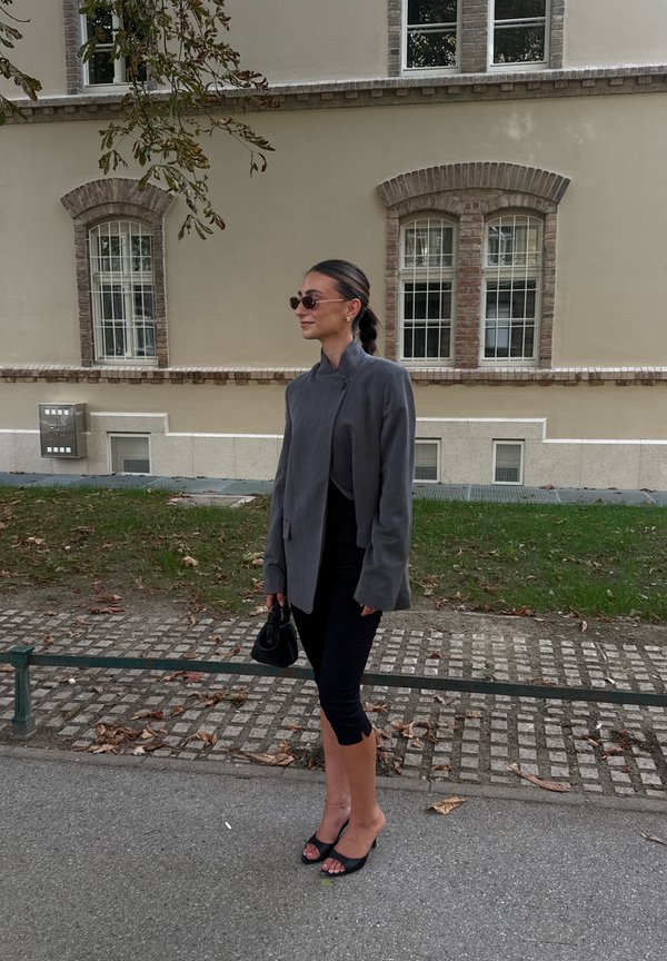 Femme portant un blazer gris, une jupe noire, des lunettes de soleil et des talons noirs, debout sur un trottoir près d'un bâtiment beige avec des fenêtres en arc.