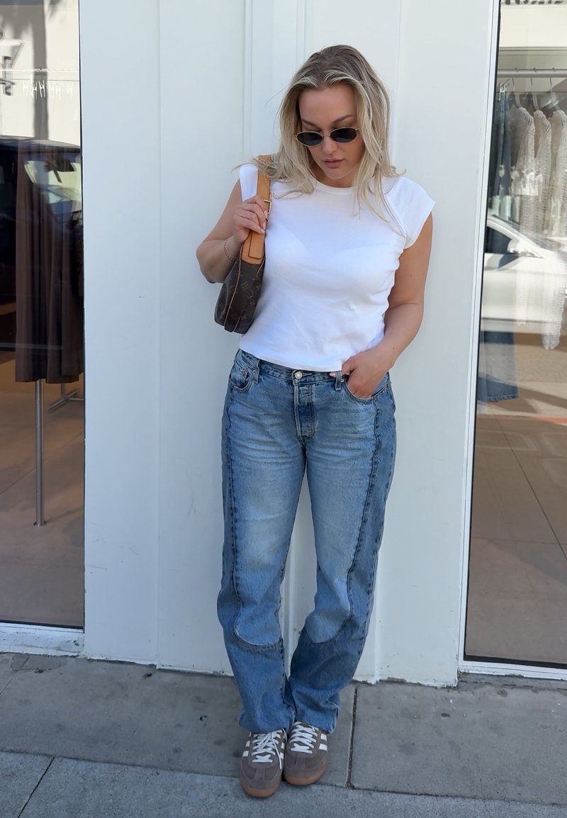 Blonde Frau mit schwarzen Sonnenbrillen, weißem ärmellosem Oberteil, blauen Jeans, grauen Turnschuhen und brauner Umhängetasche steht auf dem Bürgersteig vor einer weißen Wand.