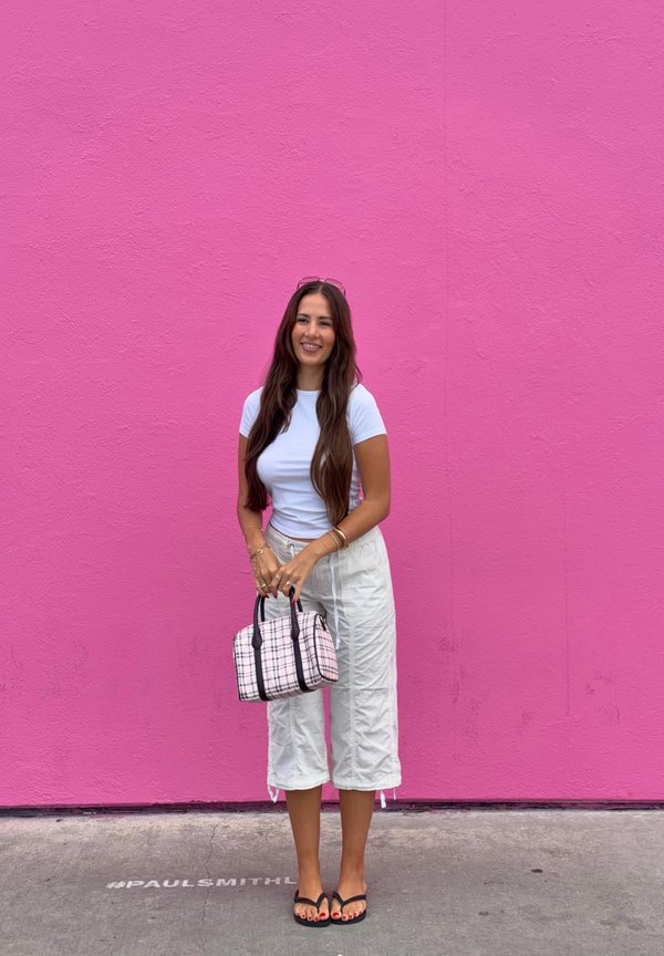 Mujer con cabello largo y castaño, vestida con camisa y pantalones blancos, sosteniendo un bolso de cuadros, parada frente a una pared rosa brillante con un hashtag en el suelo.
