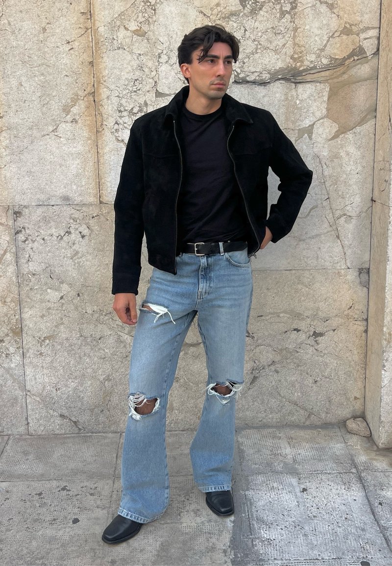 Svart semsket skinnjakke, svart t-skjorte, lyseblå slitne jeans med rifter på knærne, og svarte lærstøvler, står mot en teksturert steinvegg.