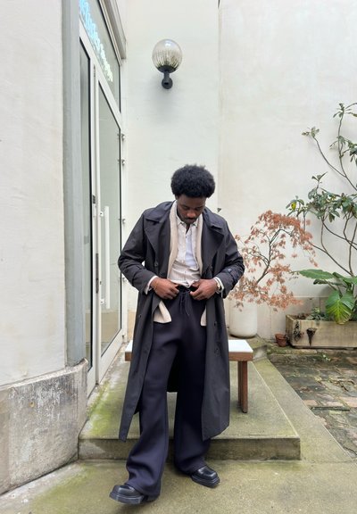 Hombre ajustándose unos pantalones oscuros y anchos, vestido con un abrigo largo negro y camisa clara, de pie al aire libre cerca de un banco y macetas contra una pared blanca.