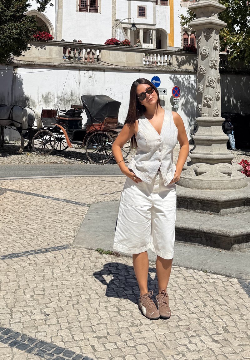 Woman with sunglasses wearing white vest and shorts stands near ornate stone pillar on cobblestone street with horse-drawn carriage in background.