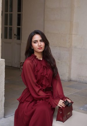 Mujer con un vestido burdeos con volantes sentada al aire libre, sosteniendo gafas de sol y un bolso a juego, con un fondo de pared y puerta de piedra.
