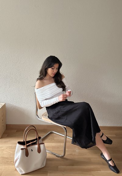 Top blanco de tirantes caídos, falda negra fluida y zapatos negros. Bolso beige con asas marrones. Suelo de madera y fondo de pared clara.