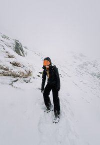 Crna vanjska jakna i hlače, narandžasta prugasta kapa, navigirajući snježnim terenom s kamenjem. Snježne pahulje padaju, maglovita pozadina.