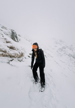 Chaqueta y pantalones negros para exteriores, gorro a rayas naranjas, navegando por un terreno nevado con rocas. Copos de nieve cayendo, fondo neblinoso.