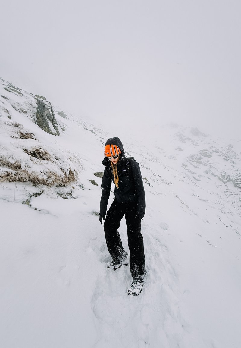 Zwarte buitenjas en broek, oranje gestreepte beanie, navigerend door besneeuwd terrein met stenen. Sneeuwvlokken vallen, mistige achtergrond.