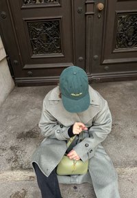 Grau aus Wolle Mantel mit einer türkisfarbenen Mütze mit goldener Stickerei; grüne Handtasche mit Logo; sitzend auf einer strukturierten grauen Oberfläche in der Nähe von verzierten Türen.