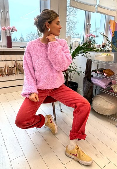 Suéter rosa y suave con mangas holgadas, combinado con un pantalón de pana rojo de talle alto y zapatillas beige con acentos rosas. Ambiente interior.