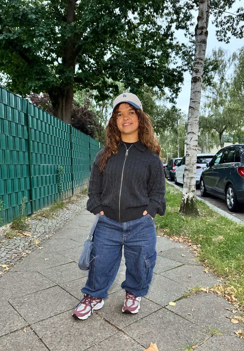 Maglione nero a maglia con zip, pantaloni cargo in denim blu larghi, cappellino grigio con logo e sneaker multicolori. Sullo sfondo ci sono una recinzione verde e alberi.