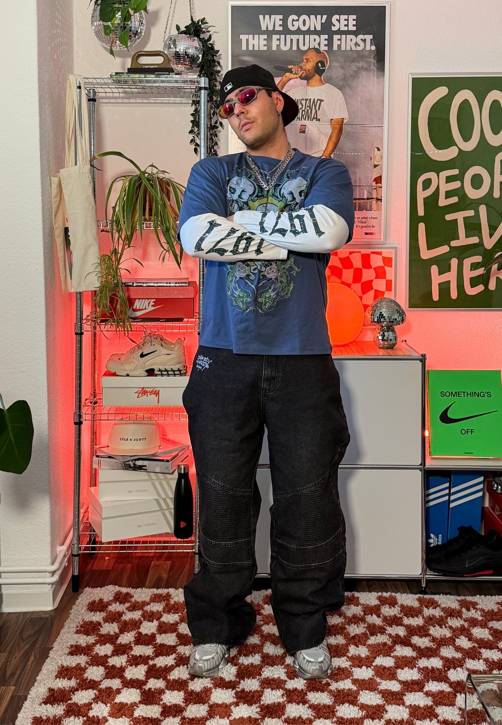 Homme portant une casquette noire, des lunettes de soleil rouges, des chemises graphiques en couches et un pantalon noir, debout les bras croisés dans une pièce vibrante avec des affiches, des plantes et des baskets.