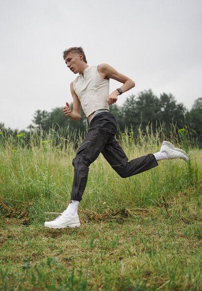 Conjunto de running que incluye una camiseta sin mangas de color beige claro, pantalones deportivos negros y zapatillas de running blancas con acentos morados en un campo de hierba.