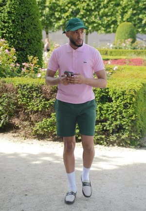 Camisa de polo rosa con un pequeño logo verde, pantalones cortos verdes, calcetines blancos y zapatos claros. Lleva una gorra verde, sostiene un teléfono, al aire libre en un jardín.