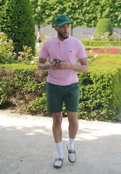 Camisa de polo rosa con un pequeño logo verde, pantalones cortos verdes, calcetines blancos y zapatos claros. Lleva una gorra verde, sostiene un teléfono, al aire libre en un jardín.