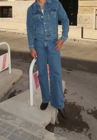Casaco de denim e jeans de boca de sino em azul claro, usado com botas de tornozelo escuras. Os acessórios incluem uma única pulseira. O fundo apresenta elementos urbanos.