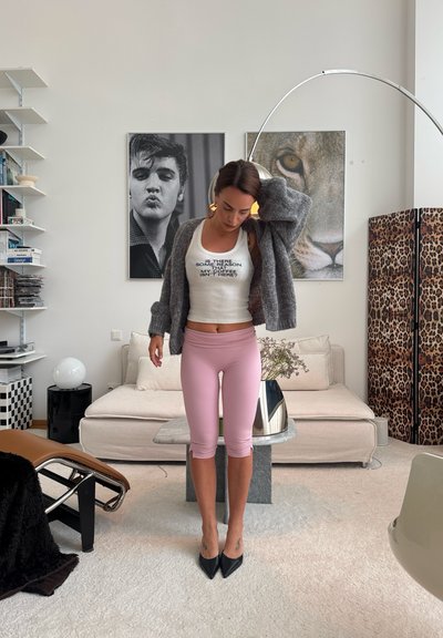Mujer ajustándose un cárdigan gris, de pie en la sala, lleva una camiseta sin mangas blanca con texto y leggings rosa, con fotos enmarcadas y pantalla de leopardo detrás.