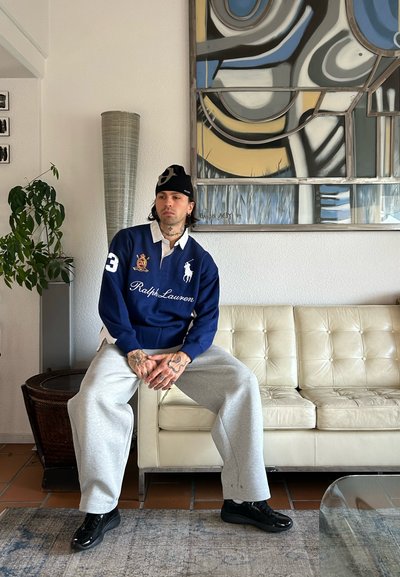 Camisa estilo rugby azul marino con cuello blanco y logo, pantalones deportivos grises, zapatos negros, sentado en un sofá de cuero blanco con una obra de arte abstracta.