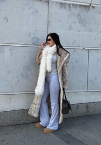 Long manteau en fausse fourrure beige porté sur un haut ajusté gris clair et un pantalon à jambes larges. Écharpe blanche texturée, sac marron et chaussons beige complètent le look.