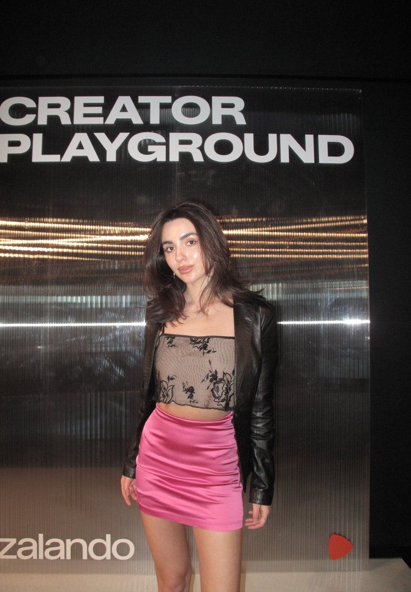 Mlada ženska, oblečena v črni cvetlični mrežasti top, črno usnjeno jakno in roza satenasto mini krilo, stoji pred zidom z napisom „CREATOR PLAYGROUND“.