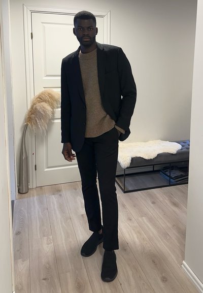 Blazer negro, suéter marrón, pantalones negros y zapatos negros; de pie sobre un suelo de madera clara cerca de una puerta blanca y un banco de felpa.