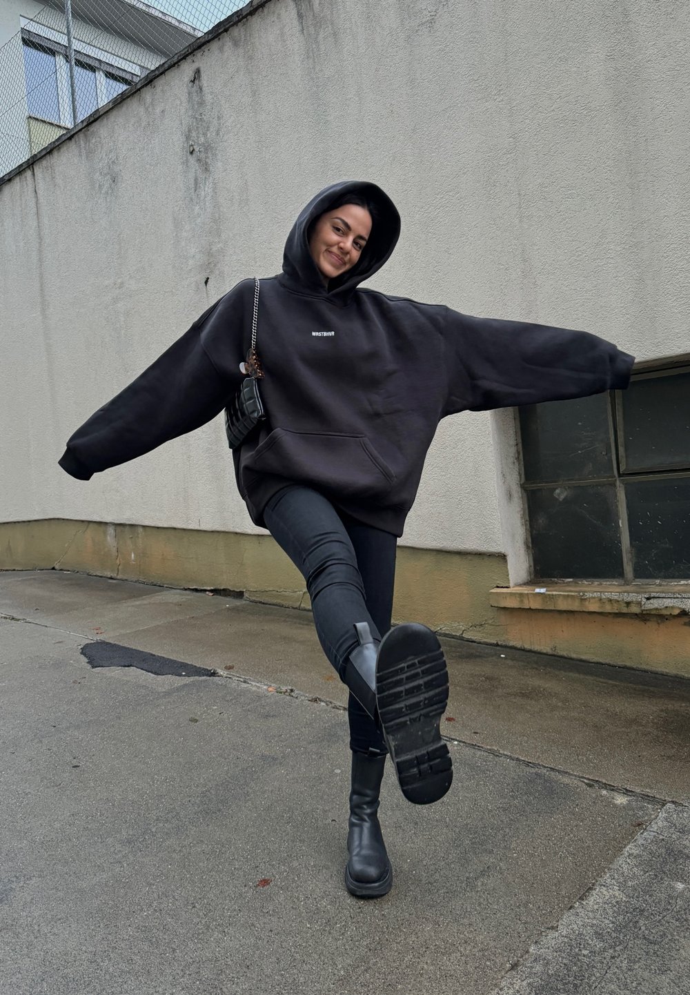 Μεγάλου μεγέθους μαύρη φούτερ με καπιτονέ τσέπη μπροστά και κουκούλα, φορεμένη με μαύρες στενές τζιν και μαύρες μπότες ύψους αστράγαλου σε φόντο από π pavement.