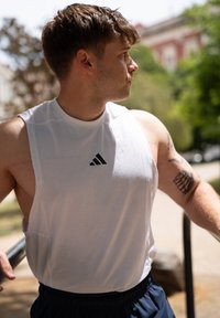Camisa de atletismo sem mangas branca com um logo Adidas preto. O modelo tem tatuagens visíveis nos braços superiores e está a usar calções escuros. Ao ar livre, sob a luz do sol.