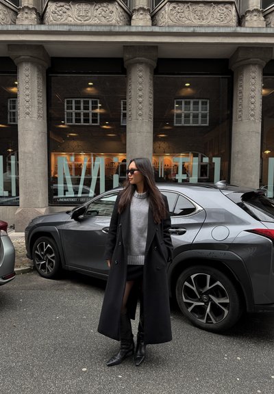 Abrigo negro de doble botonadura sobre un suéter gris, combinado con una falda mini negra y botas negras hasta la rodilla. Coche gris de fondo.