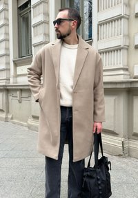 Soprabito beige in morbido misto lana, indossato sopra un maglione crema. Jeans scuri e borsa tote nera in tessuto. Texture liscia, design su misura.