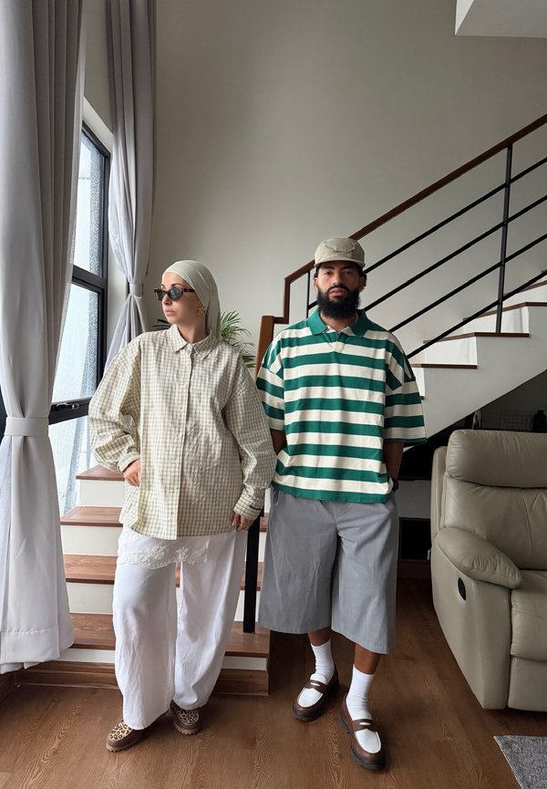 En mand i en grønstribet polo og grå shorts står ved siden af en kvinde iført en beige ternet skjorte, hvide bukser og tørklæde på hovedet nær trapper indendørs.